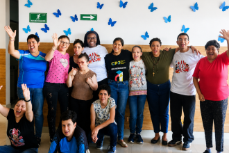 A joyful group of diverse individuals smiling and waving in front of a white wall decorated with blue butterfly decals.