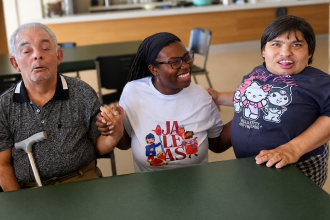 Dhani James sits at a table, laughing and holding hands with two of the center's adults.
