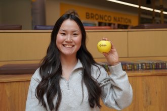 Gracie Feckovic, business education student, holds a toy apple in the student center with a sign that reads "Bronco Nation" in the background.