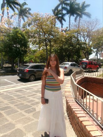 Ashley Solis is smiling, wearing a striped top and white skirt. She stands outside on a sunny day. 