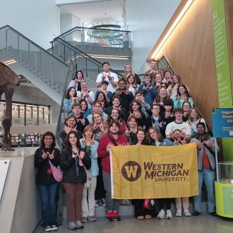 A large group of honors students stand on a staircase, the students in front are holding a WMU flag, many other students are making the W with their hands. 
