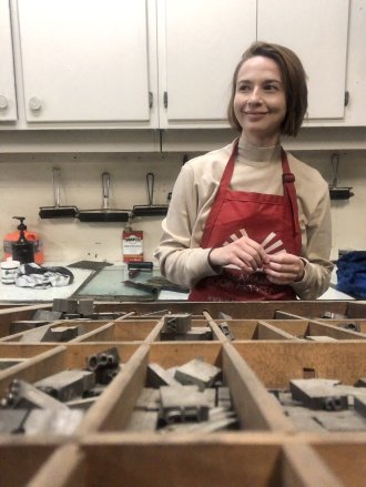 Painting alum Rachel Stickney in the studio wearing an apron behind a case of letters.