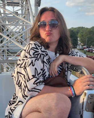 Aiden Ward sits in a ferris wheel bucket, wearing sunglasses and a patterned shirt, lips pursed.