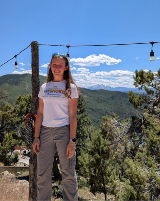 Anastasiia Hrukach stands outside, with mountains and a clear blue sky behind her, smiling. 