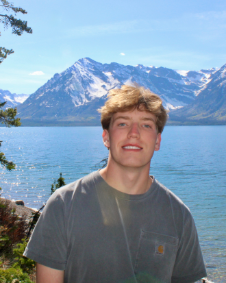 Andrew Kelly stands smiling against a brilliantly blue background of lake and mountains.