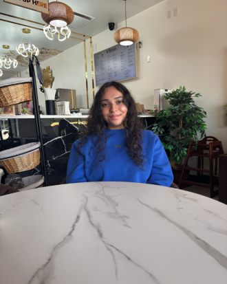 Eiman Intikhab, wearing a blue shirt, and her hair down, sits at a marble cafe table, smiling.