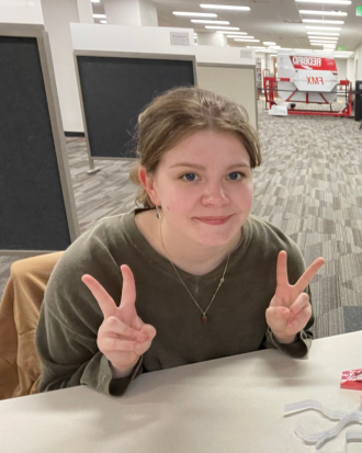 Francesca McCaughan-Perkins sits at a table in a green shirt, making peace signs with both hands, smiling.