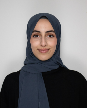 Hadeel Shebrain stands in front of a light background wearing a blue-gray headscarf, smiling. 