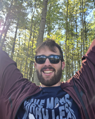 Jake Waterlander in the woods, wearing sunglasses, smiling, raising his arms in happiness.