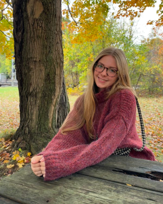 Jessica Muller, wearing a pink sweater and sitting at a picnic table, smiles. It is autumn.