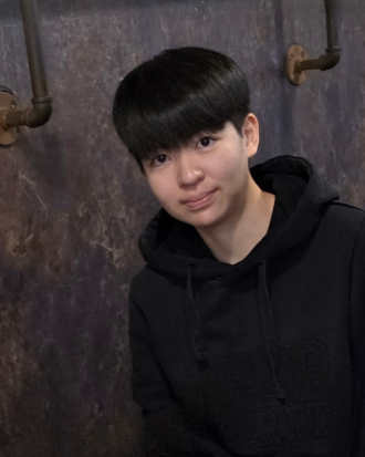 JJ Jeon, wearing a black hooded sweatshirt, stands in front of a dark wall, smiling.