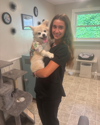 Kaylee Jangda, wearing black scrubs, stands in a tiled room holding a freshly groomed dog, smiling.