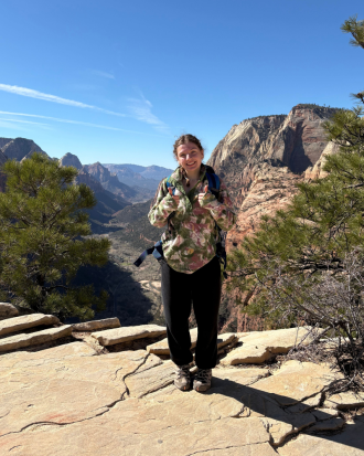 Lillian Ulmer stands on a rocky mountain top wearing a flower patterend jacket, giving the world a thumb's up, smiling.