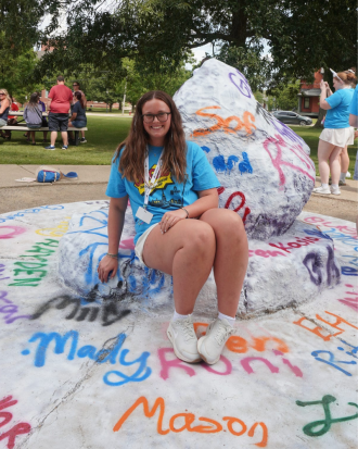 Mady Rumsey sits on a spray painted rock wearing a blue graphic t-shirt and shorts, smiling. Her name is spray painted next to her feet. 
