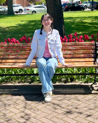 Margaret Rodgers, wearing a white jacket and blue jeans, sits on a park bench, pink tulips behind her. She is smiling, her hair is in braids.