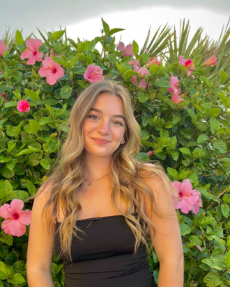 Megan Vander Kooi sits in front of a backdrop of leafy green plants spotted with pink flowers. She is smiling.