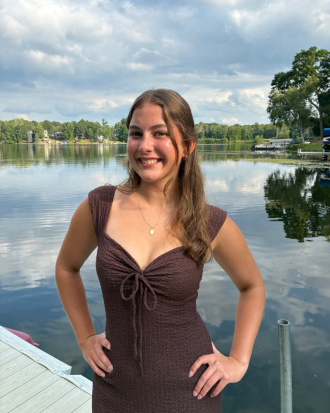 Meghan Buckwalter, wearing a brown dress, smiling, stands with hands on hips on a dock with a peaceful lake in the background. 