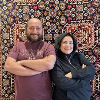 Halil and Fatma Dursunoğlu, WMU alumni and local business owners, smile together in front of a Turkish tapestry.