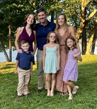 Ali Yoder, her husband and their four children pose together in front of a sunlit lakeside backdrop.
