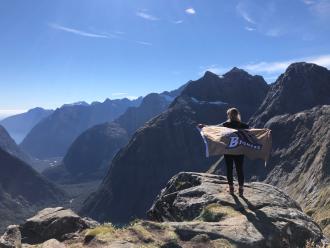 Lauren LaLonde stands at the peak of Gertrude's Saddle, overlooking Milford Sound while holding a Broncos flag behind her back