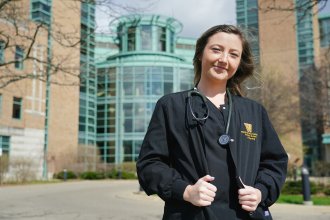 A nursing student wearing scrubs and a stethoscope stands outside Bronson Hospital.