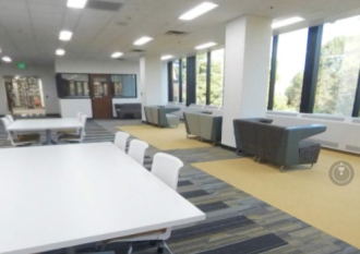 This images shows a portion of the Graduate Student Commons at Waldo Library. There is a white study table in teh foreground with three chairs on each side. A similar table is in the background on the left, and in the background in the center and on the right is a line of small sofas facing large windows with trees visible in the far distance.