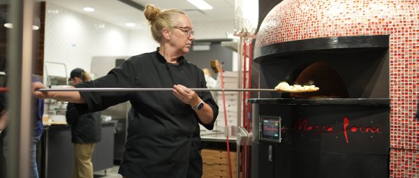 A WMU employee places a freshly-made pizza into a large red oven to bake.