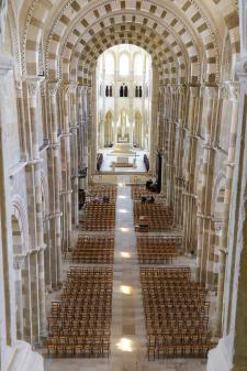Picture of the interior of a cathedral, taken from the ceiling. Arched supports line the chamber, in white and grey stone. There are rows of wooden chairs on the floor of the chamber. At the far end is the altar, carved out of white stone and lit by sunlight coming in through windows.