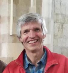 A person with short grey hair smiling at the camera, wearing a blue collared shirt and a red zippered jacket.