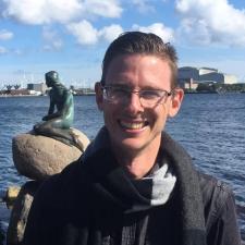 White man with short brown hair smiling, he is wearing glasses, a scarf, and a grey jacket. He is standing in front of open water, with a statue of a mermaid over his left shoulder (from the camera's perspective)..
