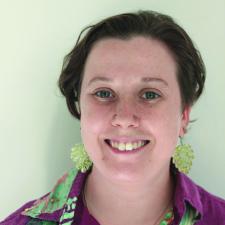 White person with short brown hair smiling, they are wearing green dangling earrings and a pink collared shirt.