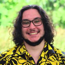 White man with long dark curly hair and facial hair smiling, he is wearing glasses and a yellow and black patterend collared shirt.