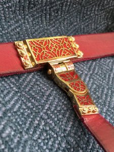 Red belt with red and gold clasp inscribed with circular motifs. 