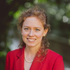 Woman wearing a red jacket and a pearl necklace; she is smiling and has curly auburn hair.