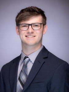 Headshot of Kevin Wright in a black suit with a grey shirt and grey patterned tie on a grey background