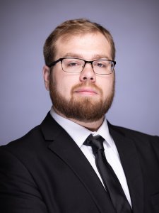 Headshot of Noah Newman in a black suit on a grey background