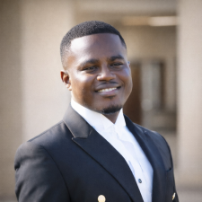 Headshot of Gabril Gyimah in a black suit with a white shirt with a hall in the background