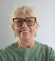 Photo of Melanie Hwalek in a green sweater and dark glasses smiling in front of a white background