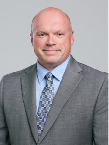 Professional headshot of a smiling bald man wearing a gray suit, light blue dress shirt, and patterned blue tie, against a light gray background.