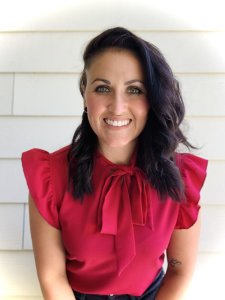 Smiling woman with long dark wavy hair wearing a bright red blouse with ruffled sleeves and a bow at the neckline, posing against a light-colored paneled wall.