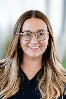 Smiling woman with long, wavy light-brown hair and patterned eyeglasses, wearing a black top and posing for a headshot against a softly blurred indoor background.