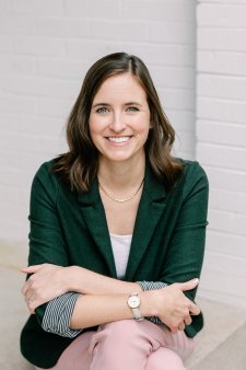 A smiling woman with shoulder-length brown hair sits with her arms crossed, wearing a dark green blazer over a white top, striped cuffs, a gold necklace, a wristwatch, and light pink pants. She faces the camera in front of a light gray brick wall.