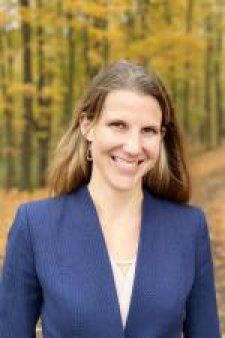 Smiling woman with long brown hair wearing a blue blazer and light-colored top, standing outdoors in front of a wooded background with autumn foliage.