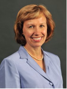 Professional headshot of a smiling woman with short auburn hair, wearing a light lavender blazer and pearl necklace, against a dark gray background.
