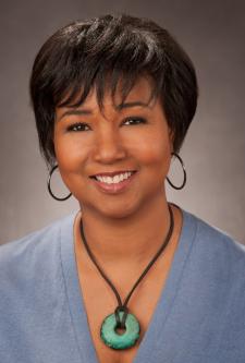 Head and shoulders portrait of a woman with short brown hair wearing a v neck top and a necklace with a large blue stone and large hoop earrings.